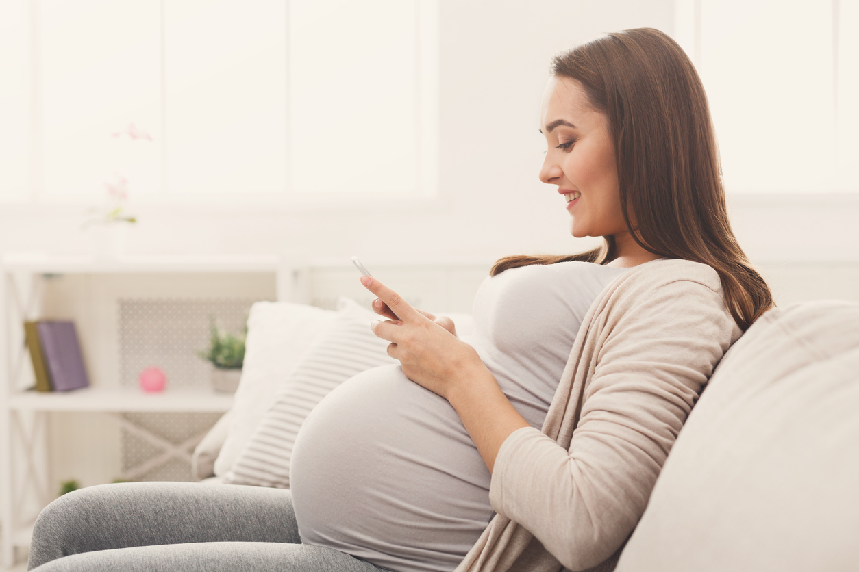 Happy pregnant woman using smartphone sitting on sofa browsing pregnancy product recommendations.