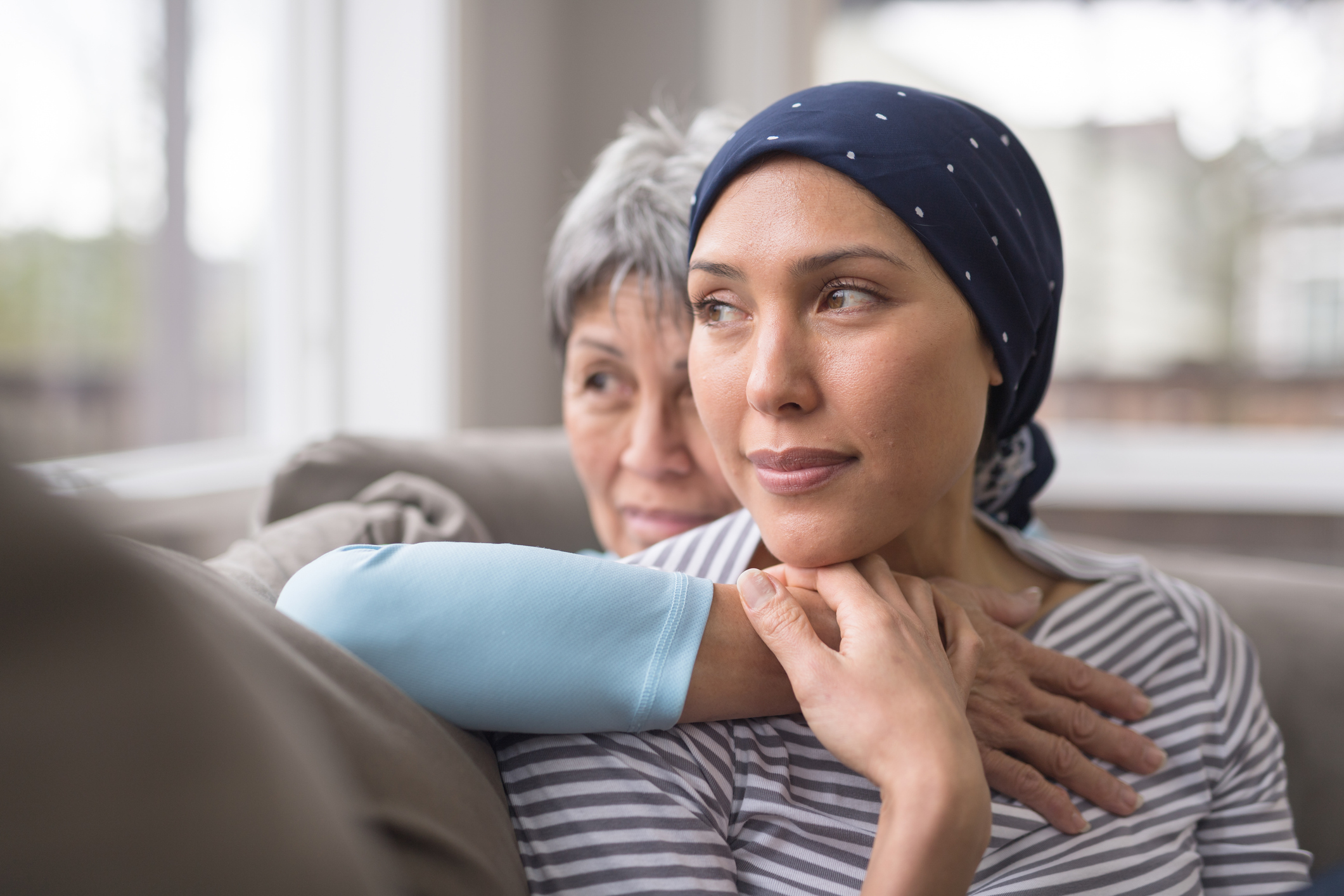 An Ashkenazi Jewish woman wearing a headscarf and fighting breast cancer sits on the couch with her mother. 